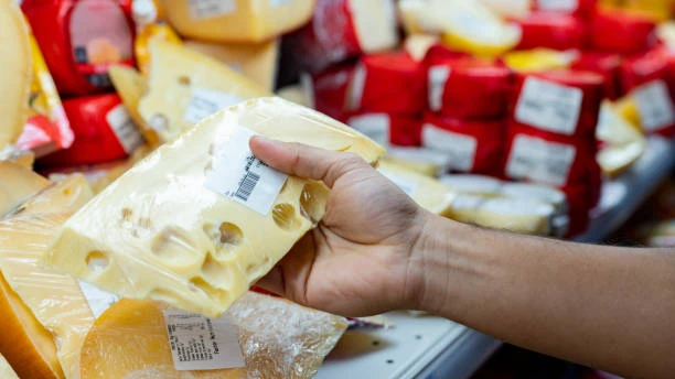 Close-up of a hand holding a vacuum-sealed chunk of Swiss cheese with characteristic holes, displayed among various cheese products in a retail grocery section